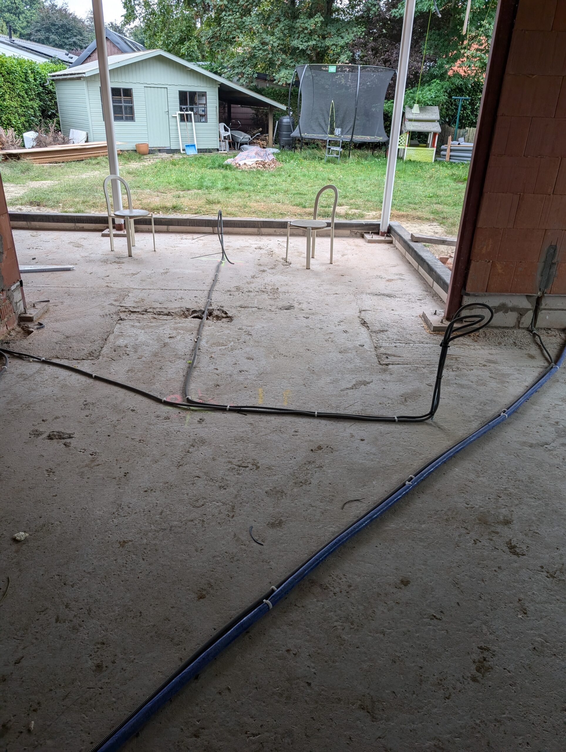 Backyard view from a partly built patio: two white metal chairs on a concrete slab with loose cables, shed and trampoline in the yard beyond.