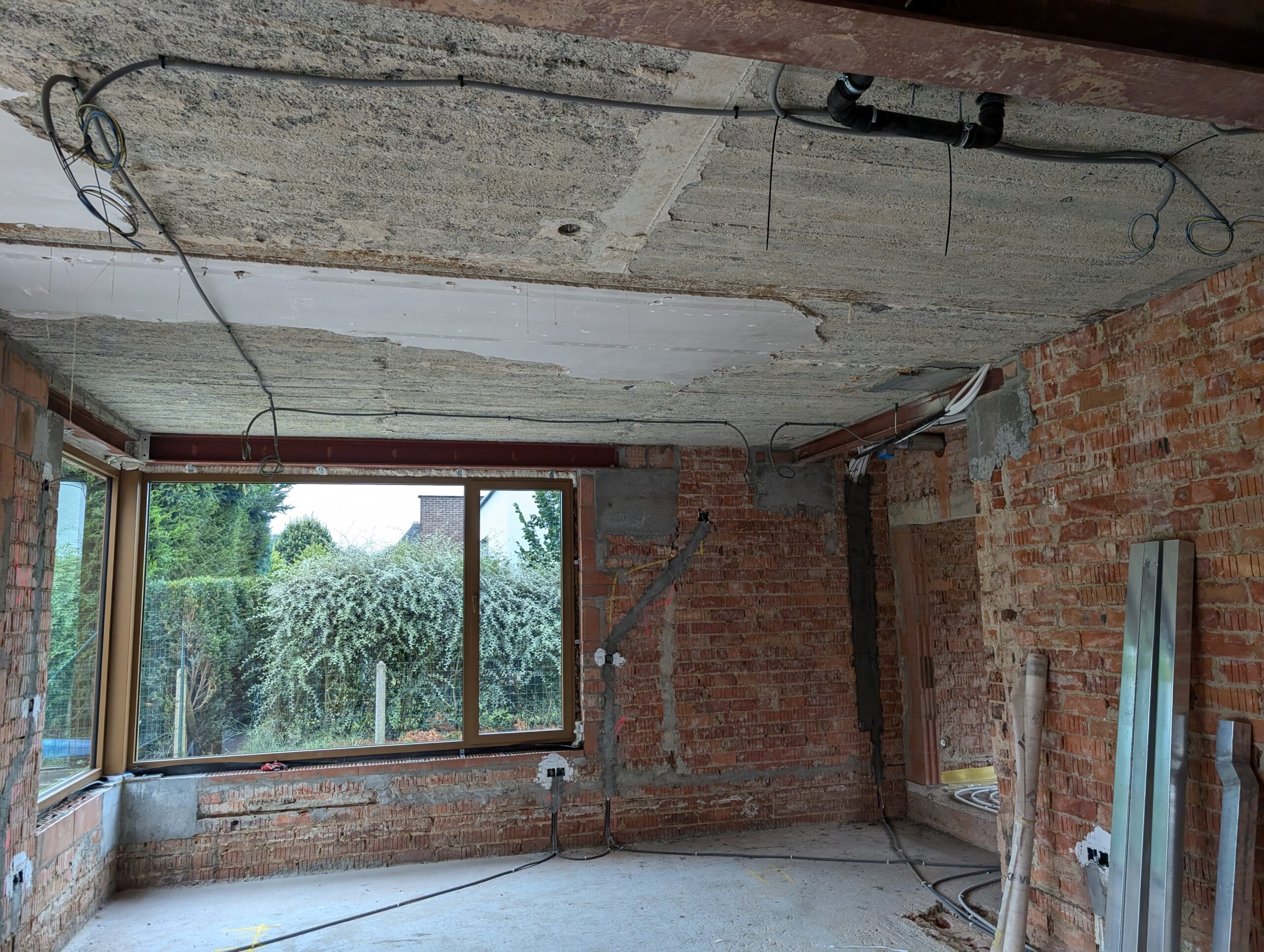 Room under renovation with exposed brick walls, a rough concrete ceiling, and loose electrical cables and pipes overhead on a construction site.