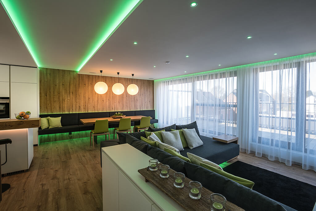 Open-concept living room with a wooden accent wall, green LED lighting, a long dark sofa with lime cushions, and sheer-curtained windows.