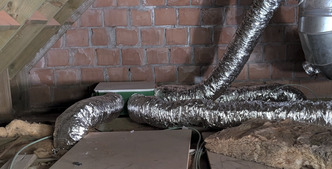 Insulated flexible foil HVAC ducts run along an attic floor in front of a brick wall, with debris and exposed insulation nearby.